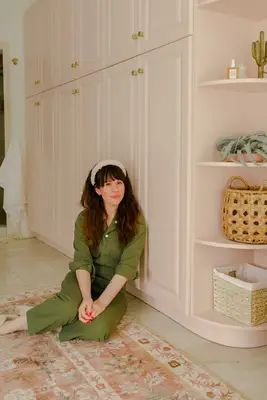 Elsie wearing green jumpsuit with a white headband sitting on the floor in front of pink cabinets