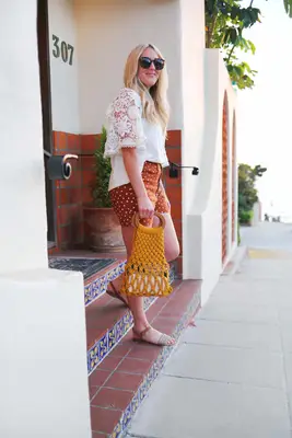a woman wearing sunglasses, a white long sleeve shirt, red with white polka dots shorts, and sandals holding a yellow macrame handbag