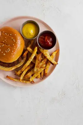 a pink plate with a hamburger, air fried seasoned french fries, a cup of mustard, and a cup of ketchup