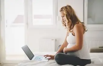 אישה בהריון יושבת על המיטה באמצעות מחשב נייד