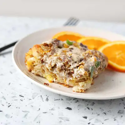 a slice of biscuit and gravy casserole and 3 orange slices on a white plate with a fork next to it