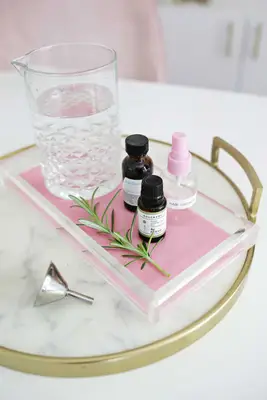 a tray with a pitcher of water, a spray bottle labeled hair growth spray, a bottle of Rosemary essential oil, and a bottle of Solubol on it with a silver funnel laying next to it