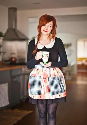 a red hair woman wearing a black dress and a flower apron with black and white checkered pockets holding a tea cup