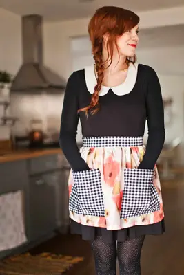 a woman with red hair wearing a black dress with a flower apron with black and white checkered pockets on