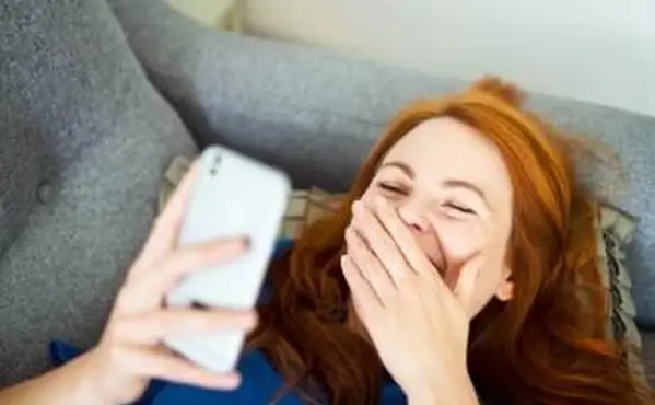 スマートフォンを使用してソファに横になって、手の後ろで笑っている女性