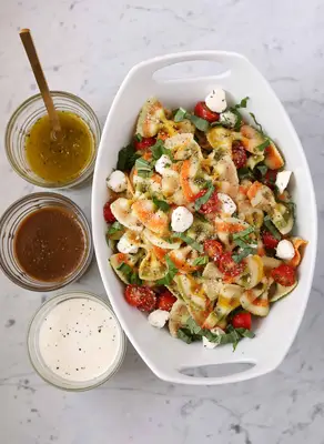 pasta salad in a white bowl with 3 small glass bowls of sauces next to it