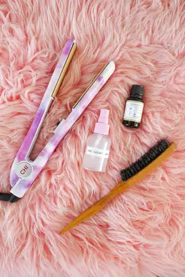 a curling iron, hair growth spray, Rosemary essential oil, and a hairbrush on a pink fluffy rug