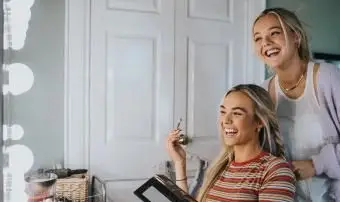 Dwie roześmiane młode kobiety przed oświetlonym lustrem i nakładają makijaż